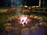 A fire burning at night time in a fire ring made of rocks.  There is a picnic table and a large pile of firewood in the background.